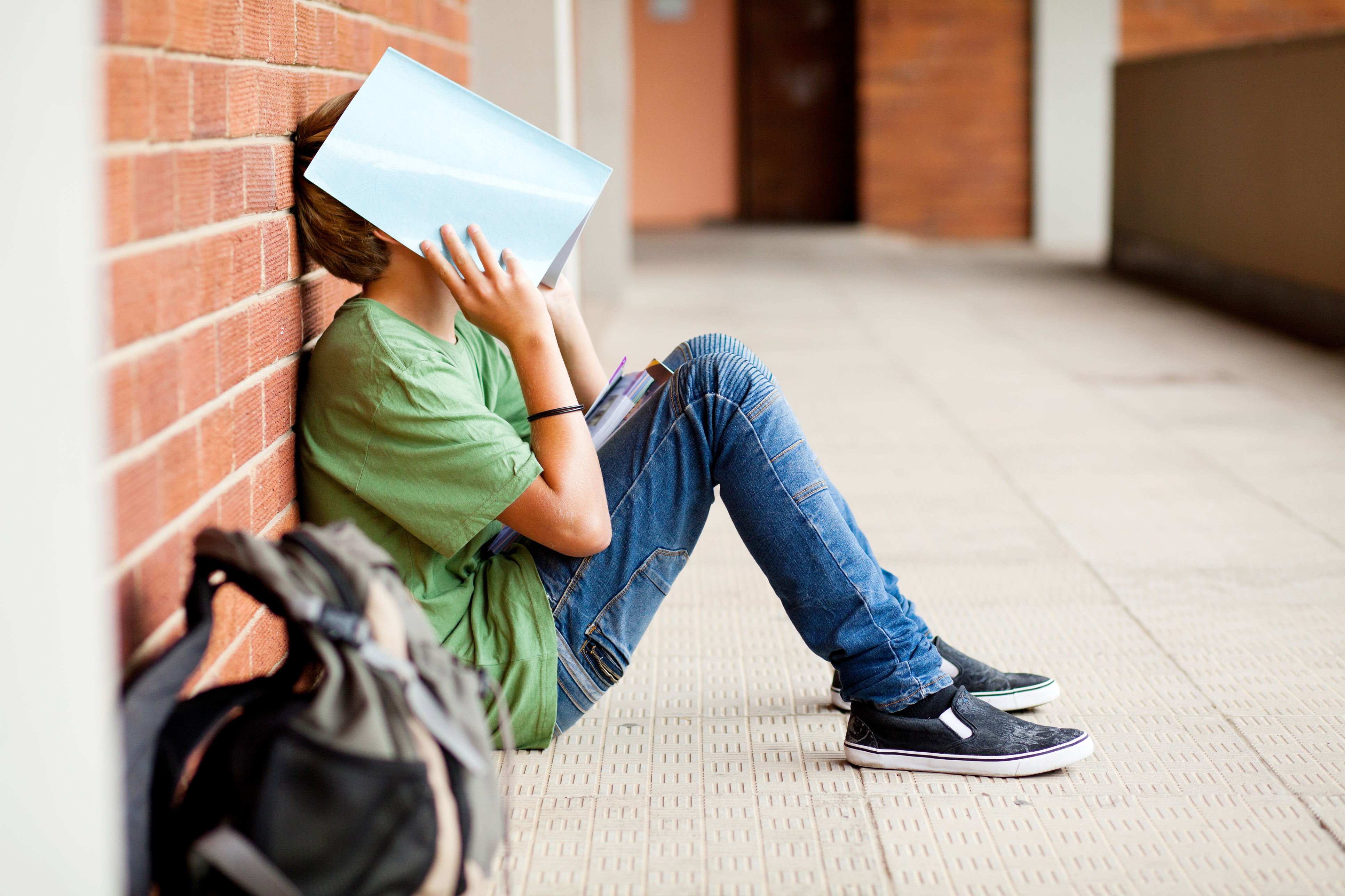 teen sitting at wall with a book over face